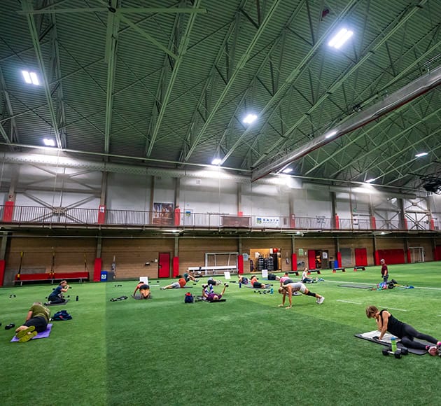 People exercising on yoga mats inside a large indoor sports facility with artificial turf.