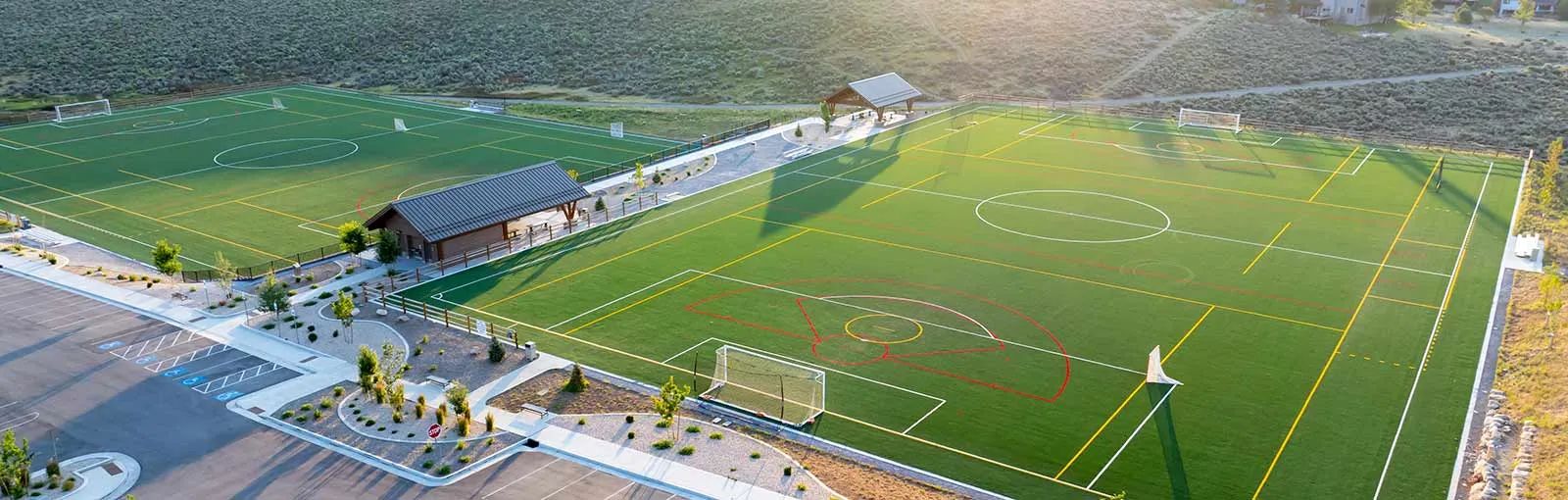 Aerial view of two artificial turf sports fields with soccer markings, surrounded by parking and landscape.