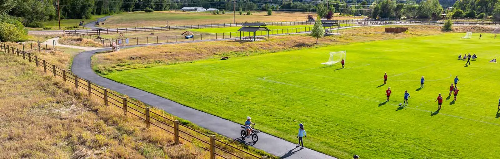 A soccer game on a grassy field with players in blue and red jerseys, bordered by a fence and a pathway with a cyclist.