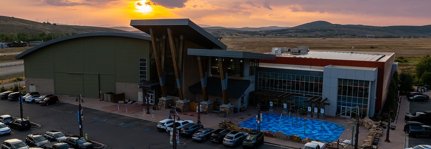 A modern building with large glass windows and an angular roof at sunset, with cars parked in front and hills in the background.