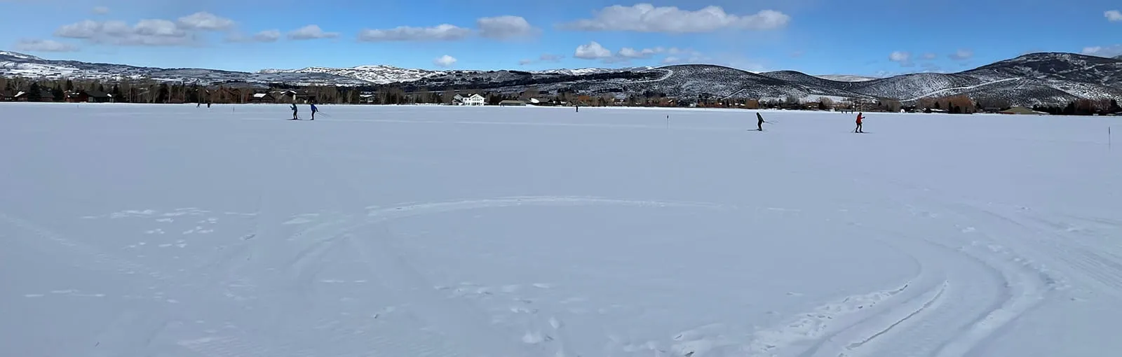 A snowy field with cross-country skiers and distant hills under a blue sky.