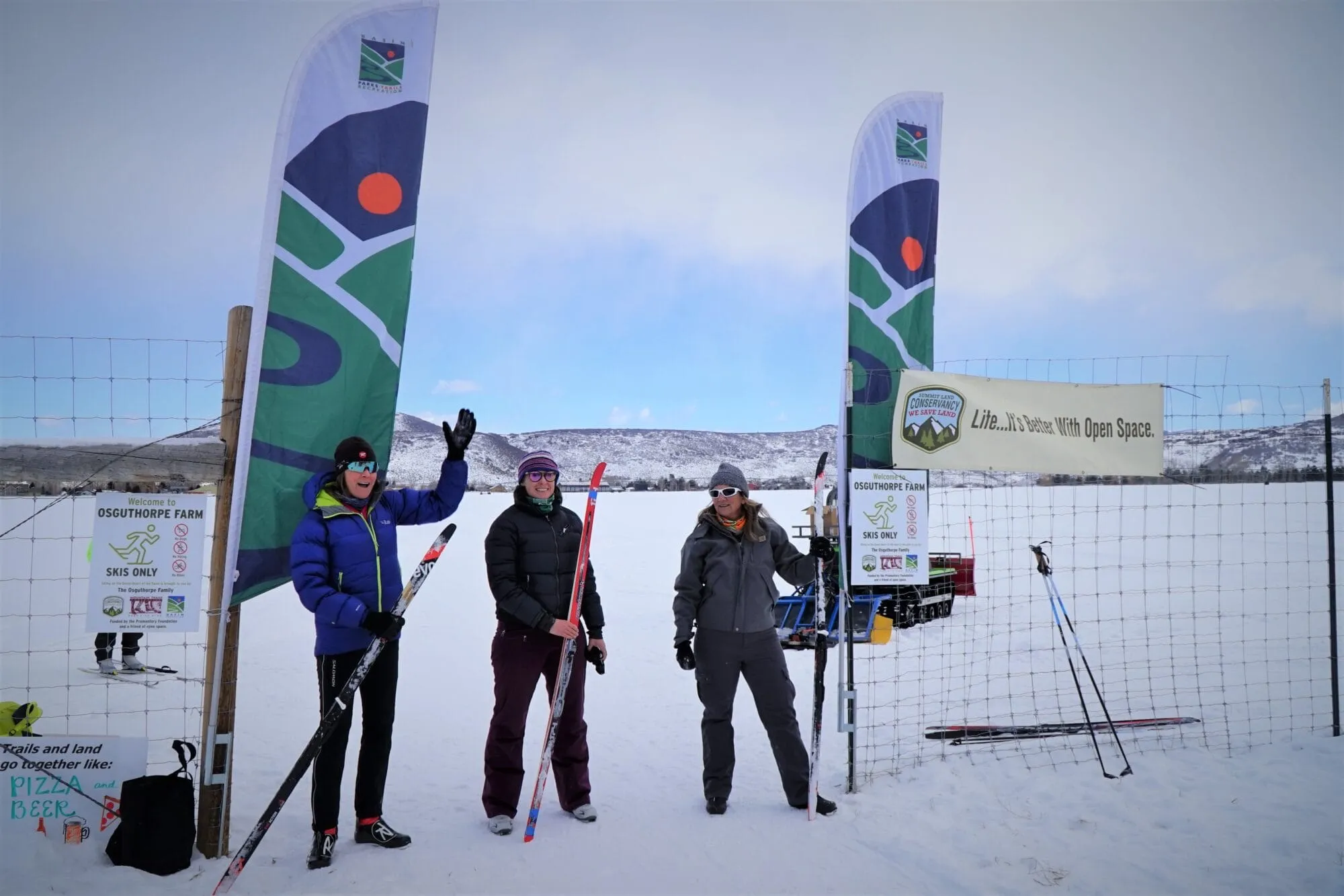 Three people stand with skis between two banner flags in a snowy field with hills in the background.