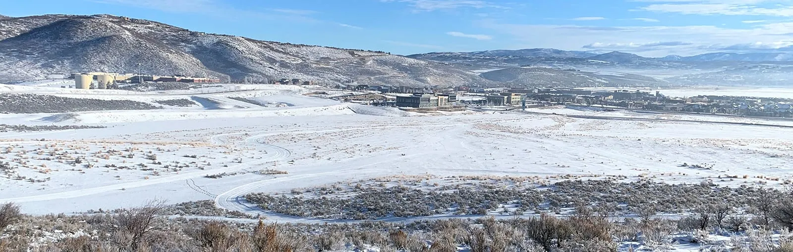 Snow-covered fields with distant buildings and snow-dusted hills under a clear sky.