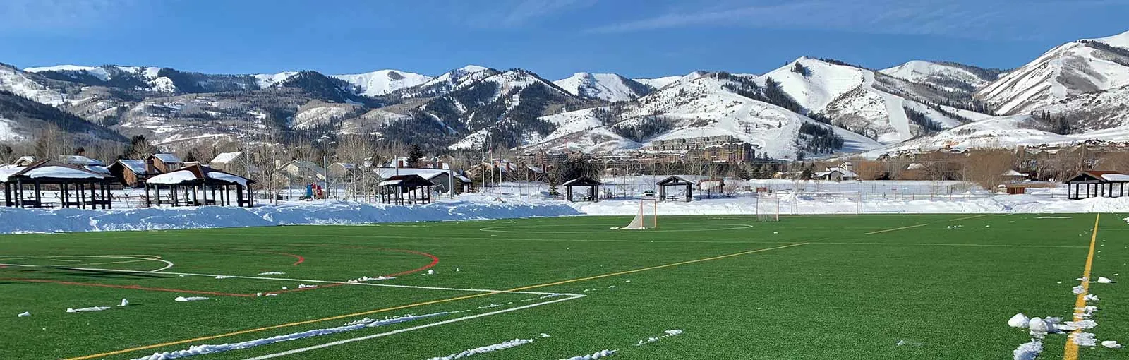 Snowy mountains behind a sports field with scattered snow.