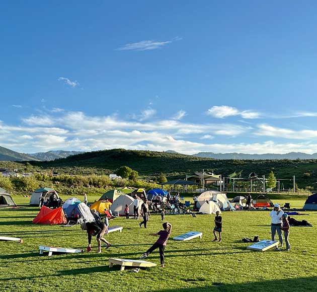 A campsite on a grassy field with tents and people playing a lawn game under a clear blue sky.