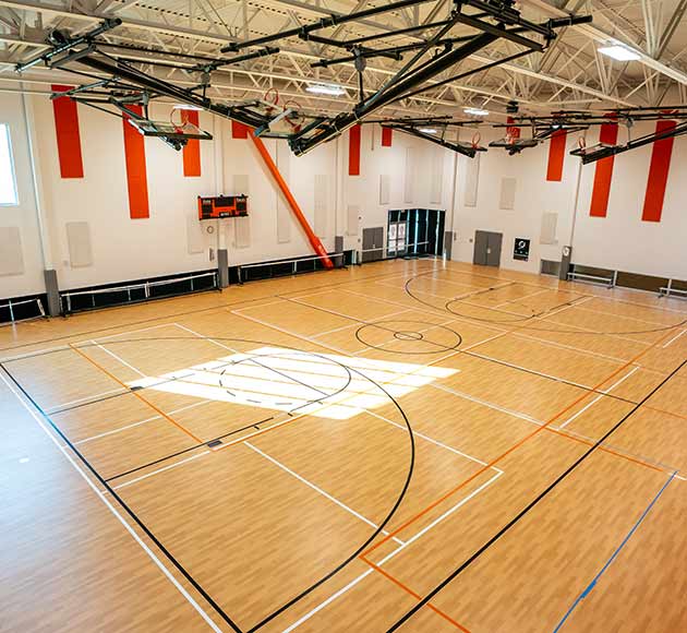 Indoor basketball court with wooden flooring, marked with black and orange lines, and retracted hoops.