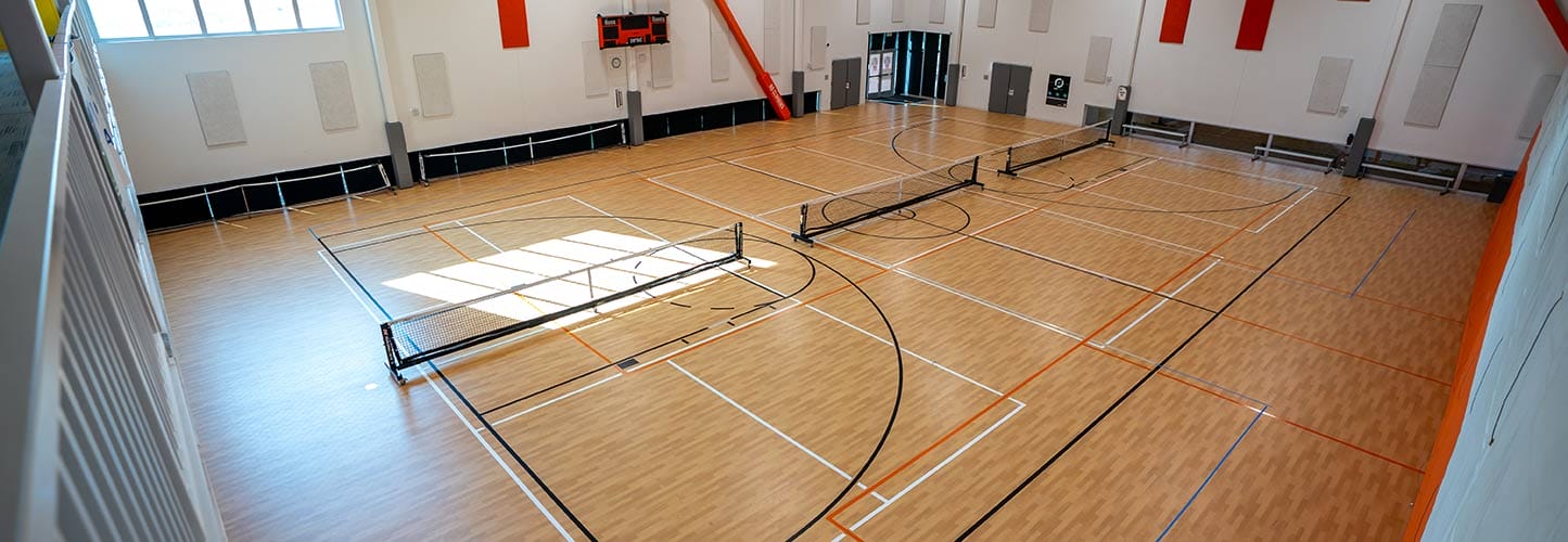 Indoor basketball court with wooden flooring, marked with black and orange lines, and retracted hoops.