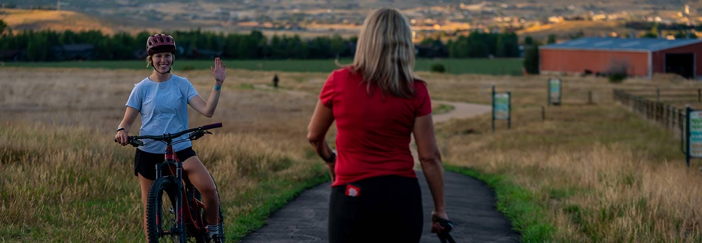 Two women on a path in a grassy field; one cycling and waving, the other walking away.