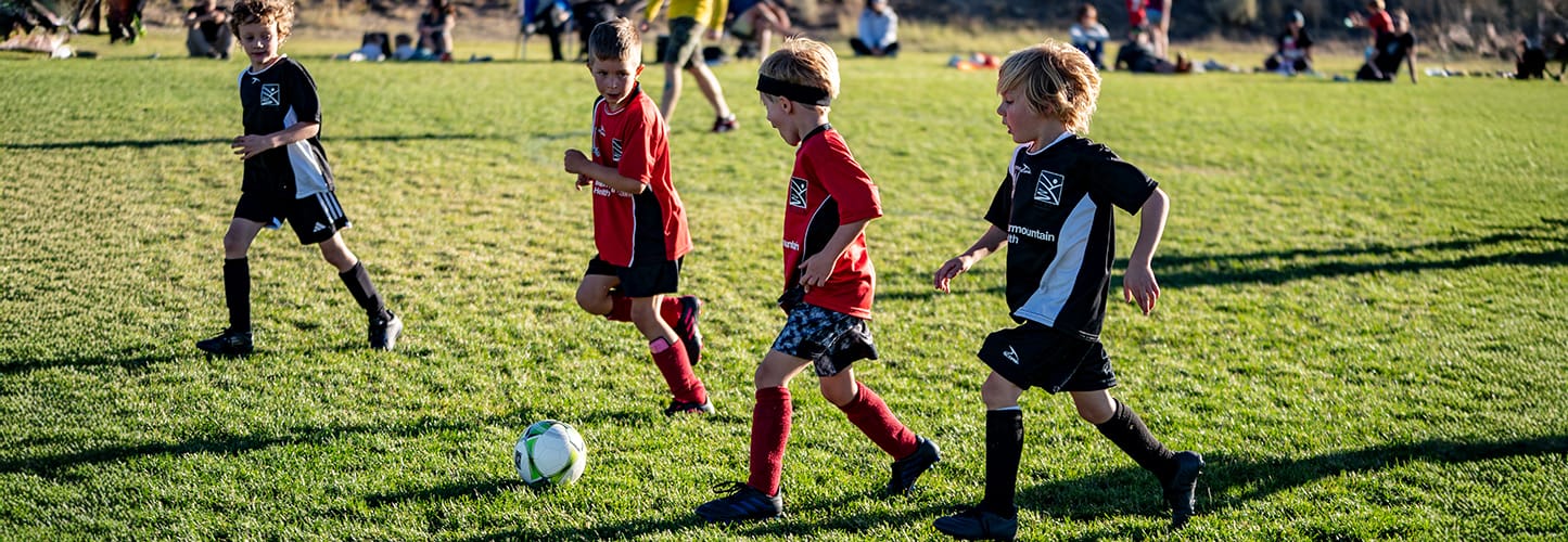 Four boys playing soccer on a grassy field, wearing red and black jerseys.