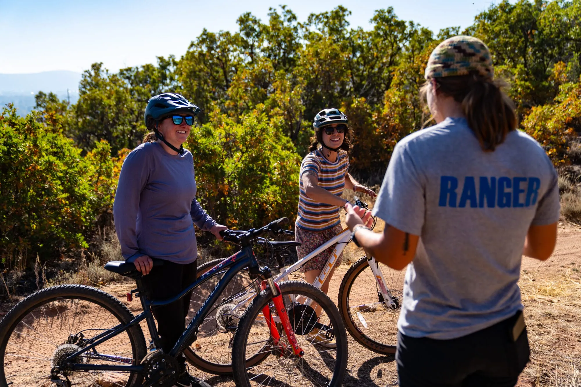 A Basin ranger standing beside two cyclists on a trail. The ranger is wearing a uniform and a hat, while the bikers are dressed in cycling gear, with bikes in the foreground.