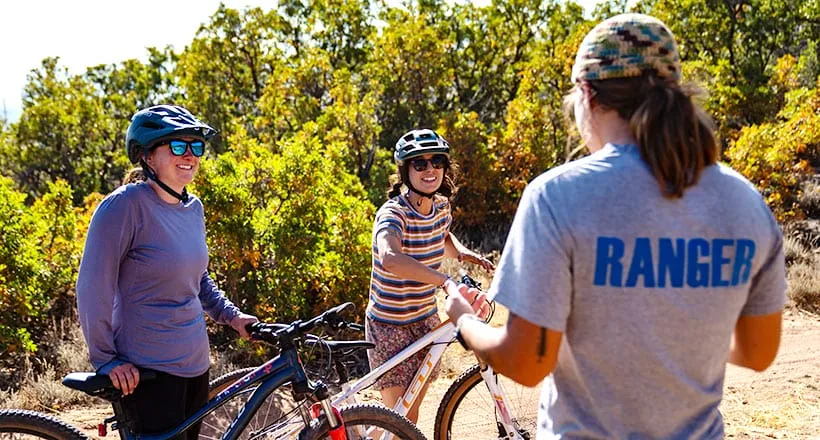 Three people conversing in a sunny, outdoor setting with bikes and greenery.