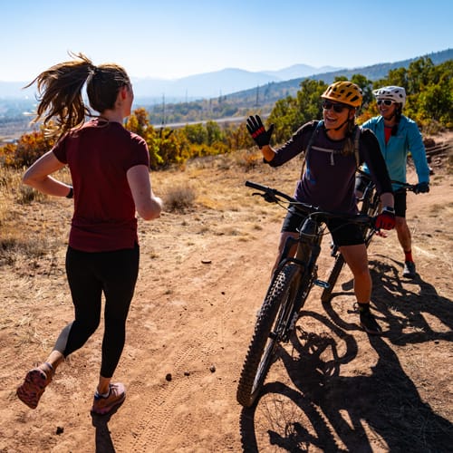 A woman running on a dirt trail gives a high-five to a cyclist wearing a yellow helmet, with another cyclist nearby under a sunny, mountainous backdrop.