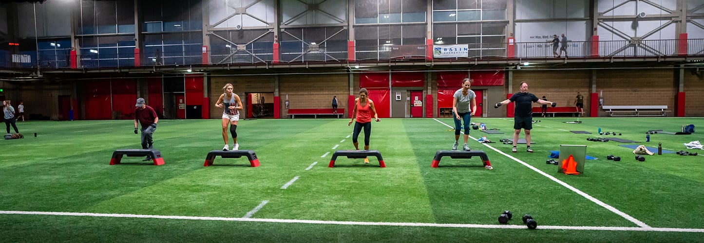 People exercising on a turf field with step platforms and dumbbells in an indoor sports facility.