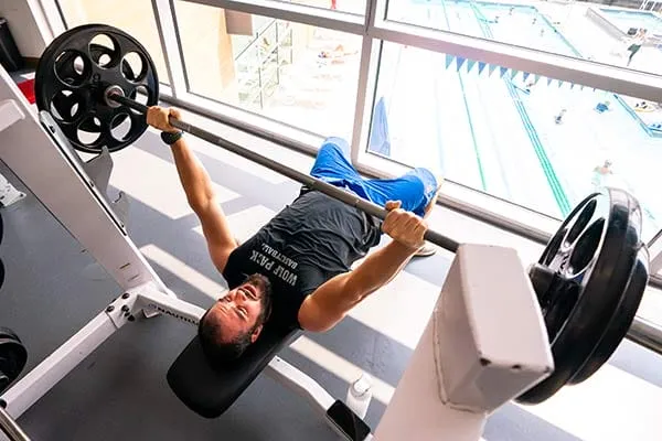 Person bench pressing in a gym near large windows with a swimming pool view.