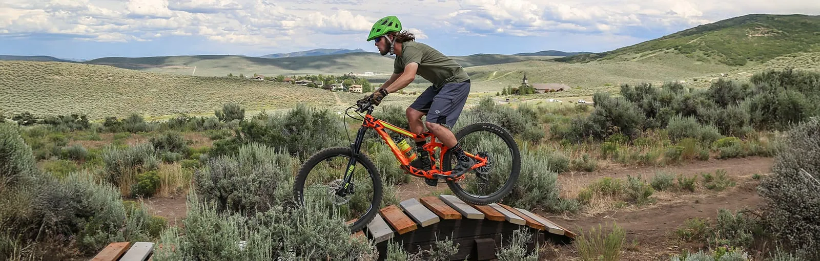 A person riding a mountain bike over a wooden ramp in a hilly landscape.