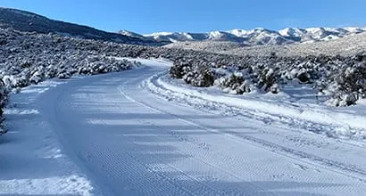 Snow-covered landscape with a groomed path and distant mountains under a blue sky.