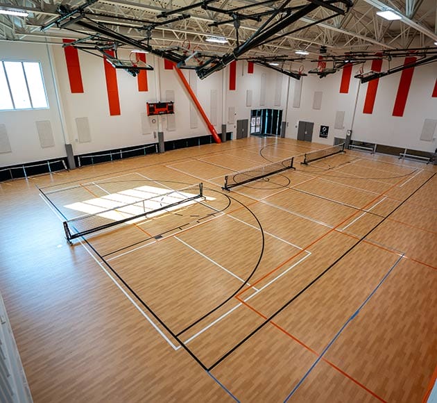 Indoor basketball court with wooden flooring, marked with black and orange lines, and retracted hoops.