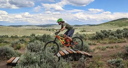 Cyclist on a mountain bike navigating a ramp in a grassy landscape under a blue sky.